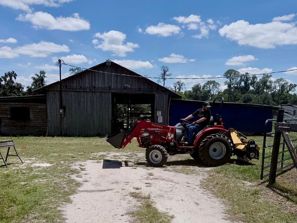 Ryan Hostetler on a compact tractor with a front loader and a rear-mounted cutter, beside the barn on the Dover acreage